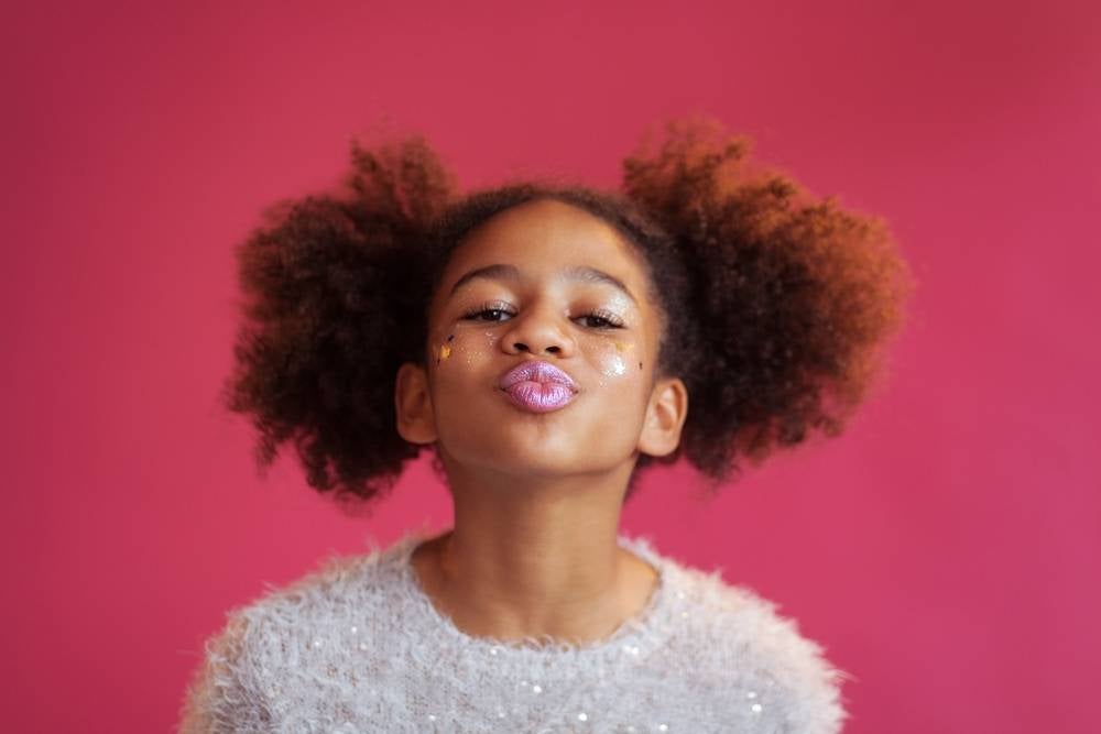 Foto de uma menina usando maquiagem colorida e uma blusa felpuda fazendo bico com a boca sobre fundo rosa. 