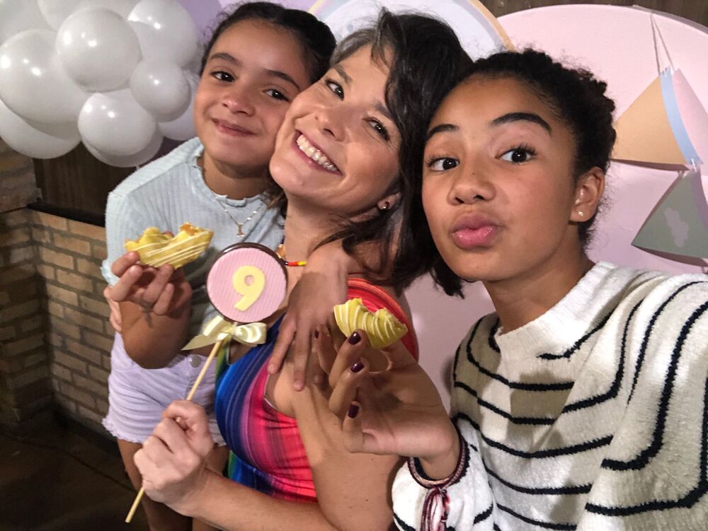 Samara Felippo e suas duas filhas em ambiente de festa de aniversário, segurando docinhos nas mãos e sorrindo para a fotografia.