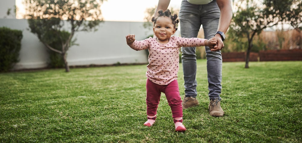 Imagem de um bebê cuja criação segue a abordagem de Emmi Pikler e que está brincando em um quintal supervisionado pela mãe que está lhe dando a mão.