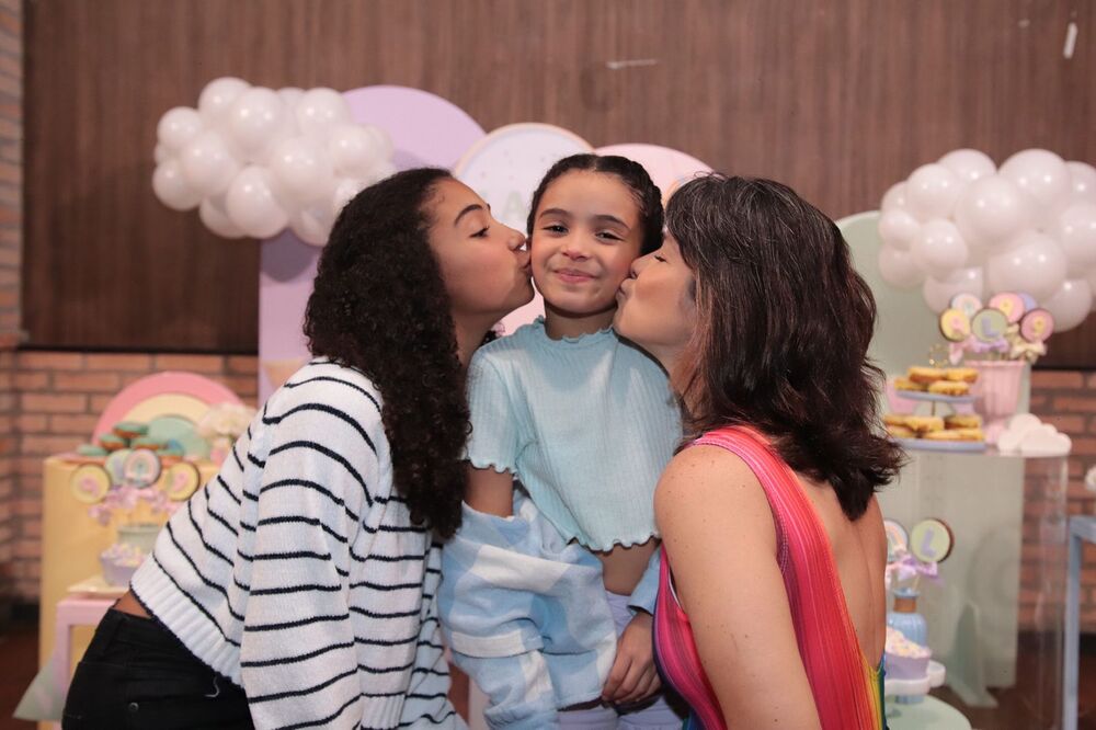 Samara Felippo e suas duas filhas. A filha mais nova no meio, mãe e irmã beijando suas bochechas uma de cada lado. ao fundo, decoração de aniversário.