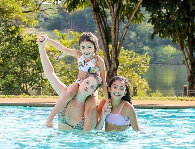  Samara Felippo na piscina com suas duas filhas, todas sorrindo em um dia de sol.