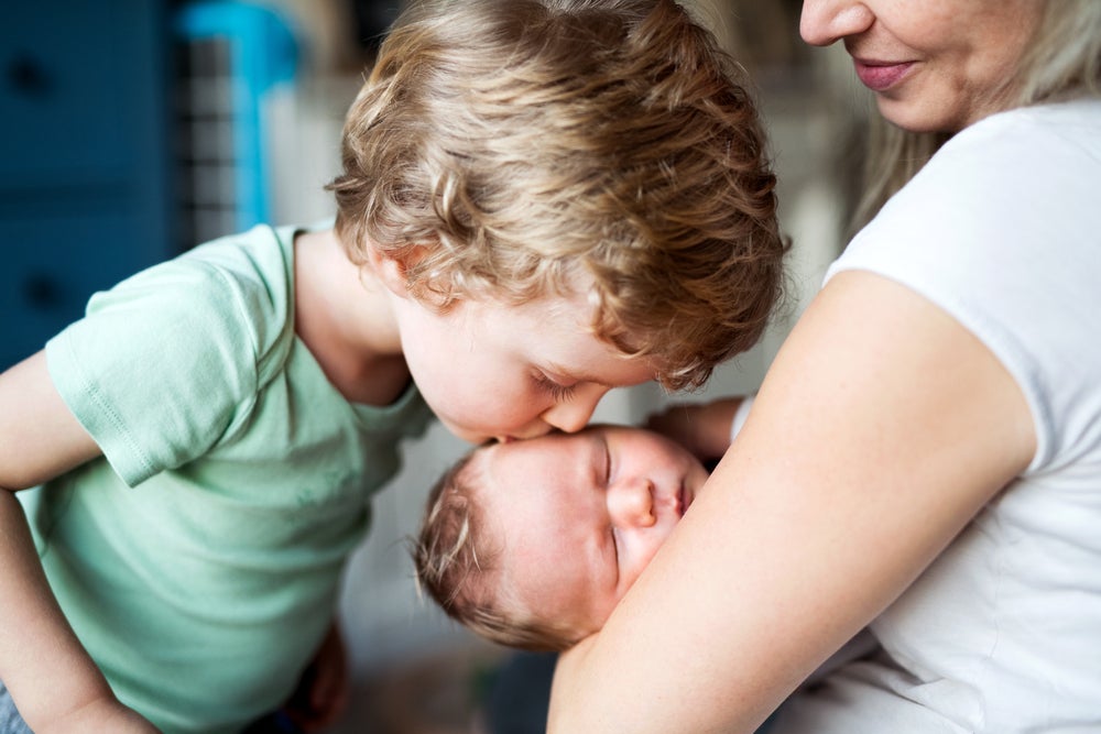 a foto mostra uma criança beijando a cabeça de um bebê que está no colo de uma mulher