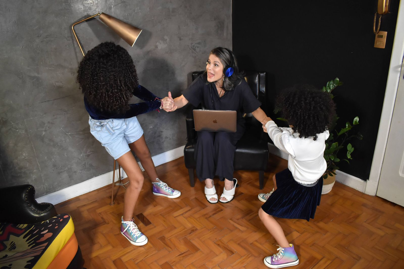 Samara Felippo com as suas duas filhas estão em uma sala. Samara está sentanda em um poltorna com o notebook no colo enquanto suas filhas estão puxando os braços da mãe para levantá-la