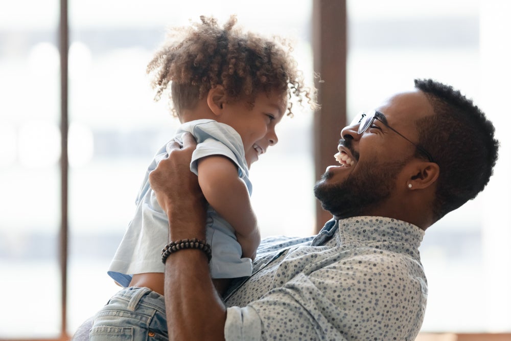 Uma criança é carregada pelo seu pai. Os dois estão brincando e sorrindo. A imagem remete ao tema crise dos três anos