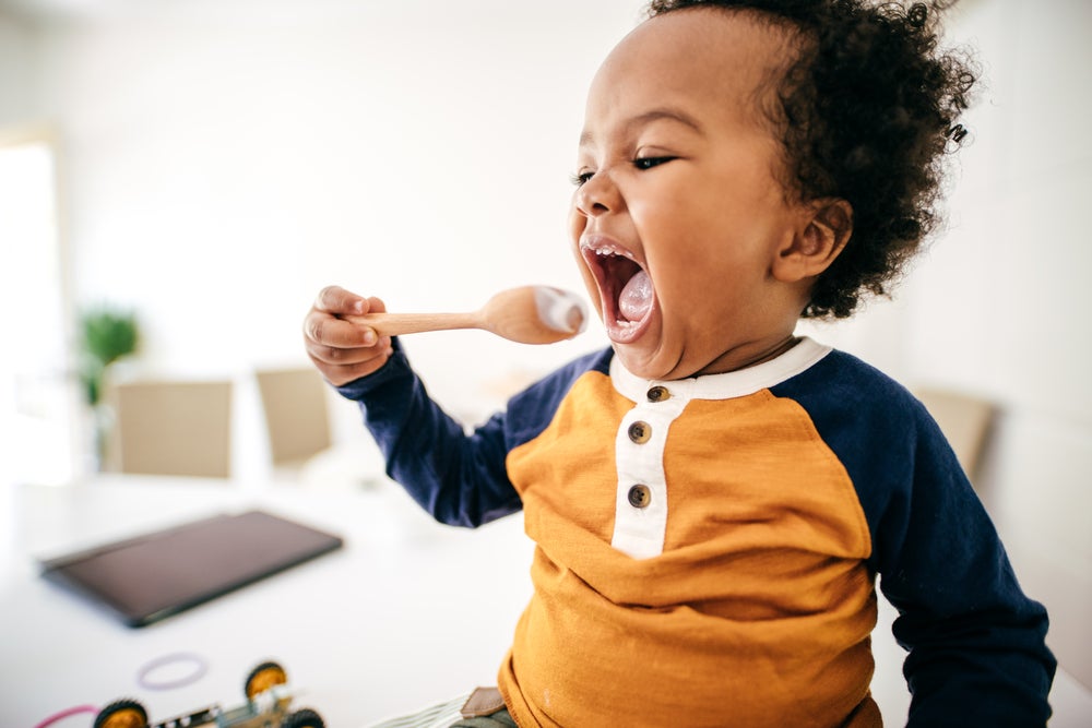 Na cozinha de uma residência temos a imagem de um bebê segurando uma colher, com a boca bem aberta, se alimentando. 