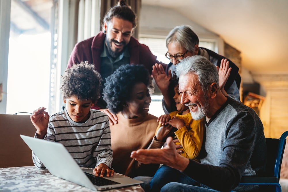 Imagem de uma família reunida na sala, olhando com felicidade a tela de um notebook. Ela é composta por um casal jovem e duas crianças, além de outros dois membros que são os avós dessas crianças. A foto remete ao tema rede de apoio materno