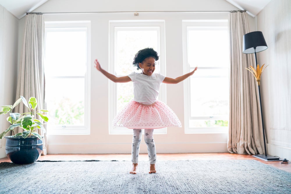 Menina vestindo um tutu de balé no meio da sala de casa. Ela se encontra com um semblante feliz e dançando pelo espaço. A foto faz alusão ao tema ballet infantil.