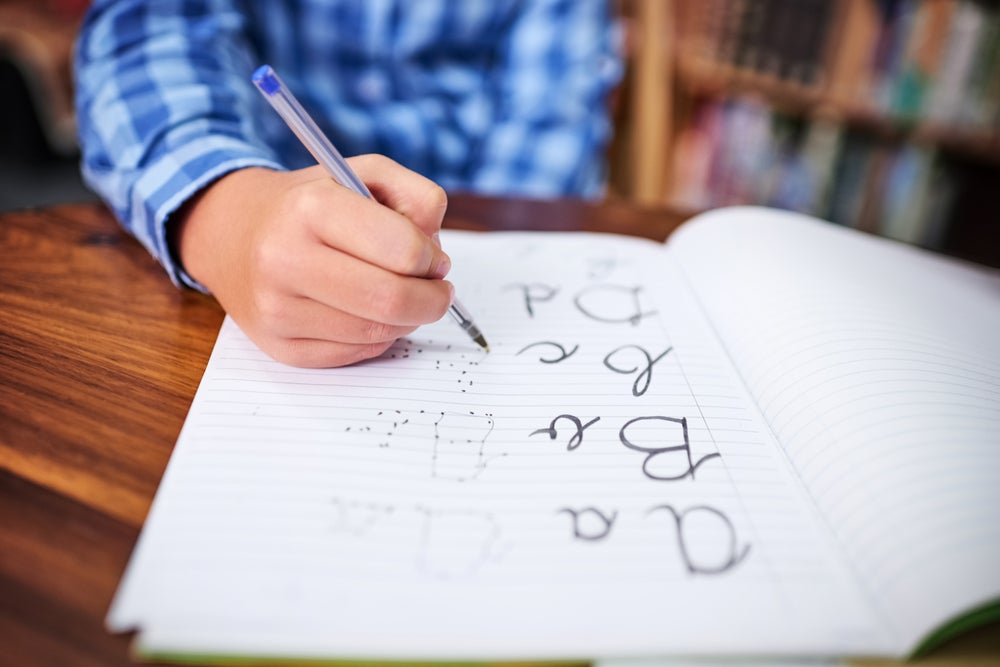 Imagem de um menino segurando uma caneta azul. Em um caderno ele vai ligando os pontinhos para formar as palavras: a,b,c e d. A foto faz alusão ao tema caligrafia.