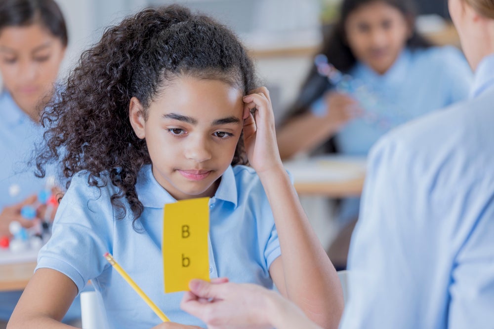 Imagem de uma criança com dislexia olhando para uma ficha amarela com letras B maiúscula e minúscula. Em uma mão ela segura um lápis e a outra está sobre a cabeça em uma postura de dúvida. 
