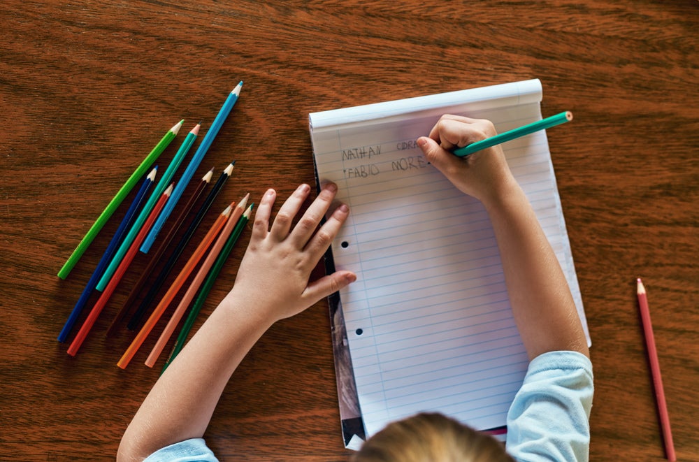 Um menino está escrevendo em um bloco de folhas alguns nomes, como “Fabio”. Na mesa estão espalhados lápis de cor de diversas cores. A foto está ligada ao tema Troca de letras na infância.