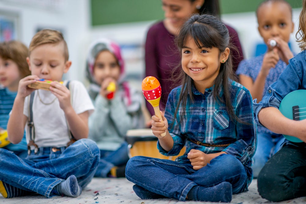 Em uma sala de aula temos algumas crianças participando de uma atividade com músicas. Em destaque, uma menina sentada no chão segurando um instrumento musical que lembra um chocalho. A imagem remete ao tema musicalização