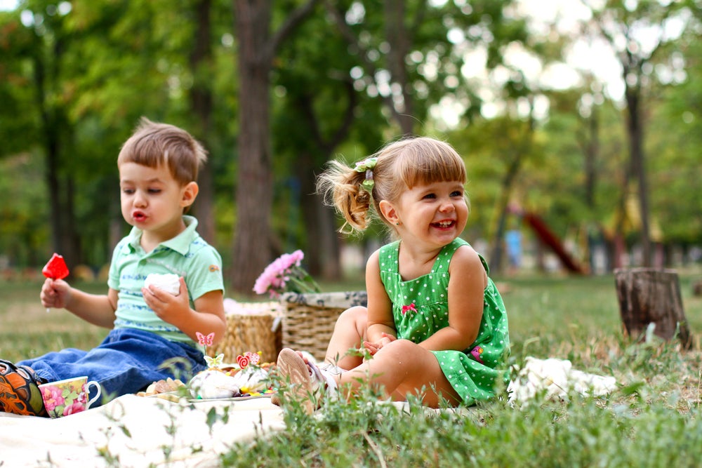 Um menino e uma menina estão sentados em uma toalha em um parque. Sobre ela temos algumas cestas e lanchinhos. O cenário todo remete muito a um dia de piquenique