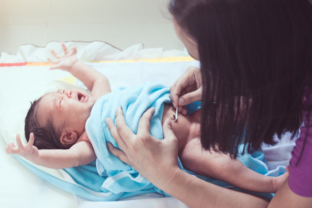 Imagem de um bebê deitado, envolvido em uma manta azul, chorando, enquanto a cuidadora limpa seu coto umbilical com um cotonete