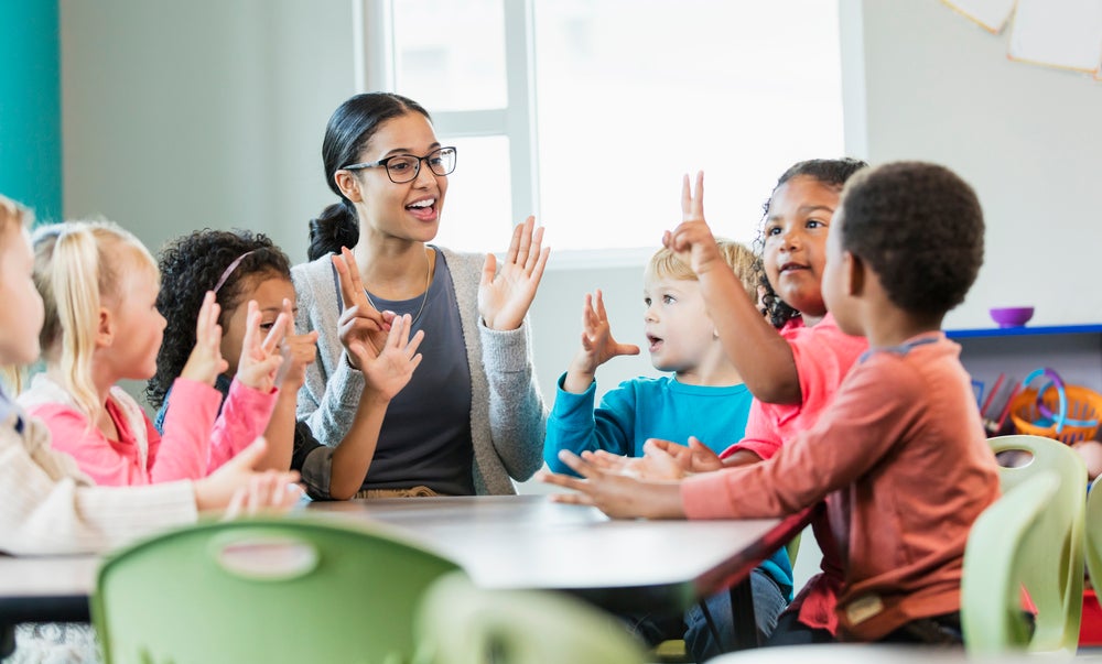 Imagem de uma tutora interagindo com crianças ao redor de uma mesa. Brincadeiras entre professores e crianças ajudam na adaptação na creche dos pequenos