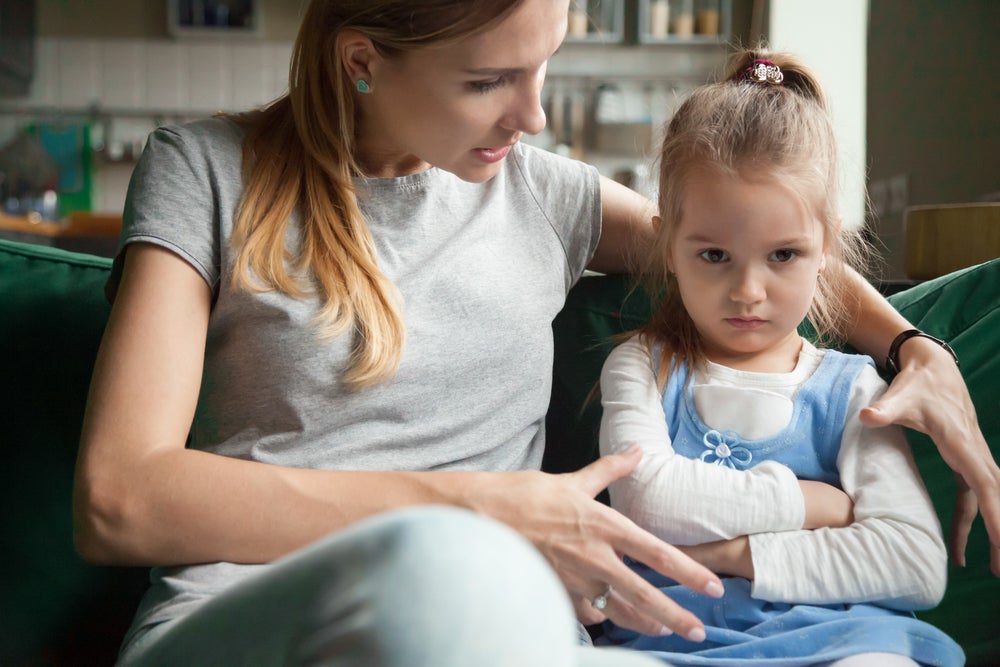 mãe e filha sentadas no sofá da sala, mãe abraça a criança e conversa, criança parece estressada com os braços cruzados e olhar distante