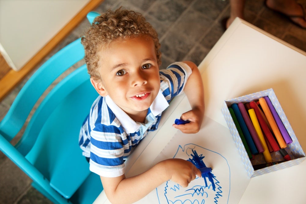criança sentada em cadeira e mesa infantil com giz de cera azul em mão, desenhando em folha de papel, outras cores de giz disponíveis sobre a mesa