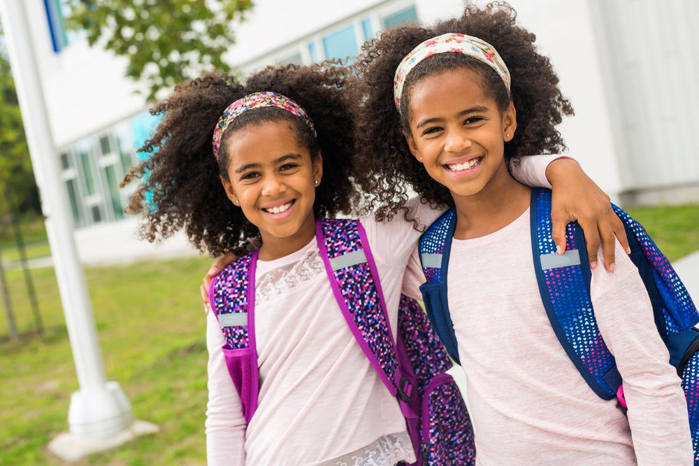 Duas irmãs gêmeas se abraçando lado a lado, sorridentes, com mochila nas costas, ao ar livre em frente a escola.