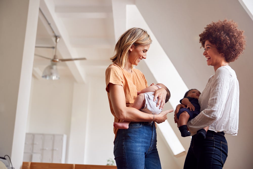 Duas mulheres adultas estão carregando seus respectivos bebês no colo, enquanto conversam, em um ambiente interno de uma residência