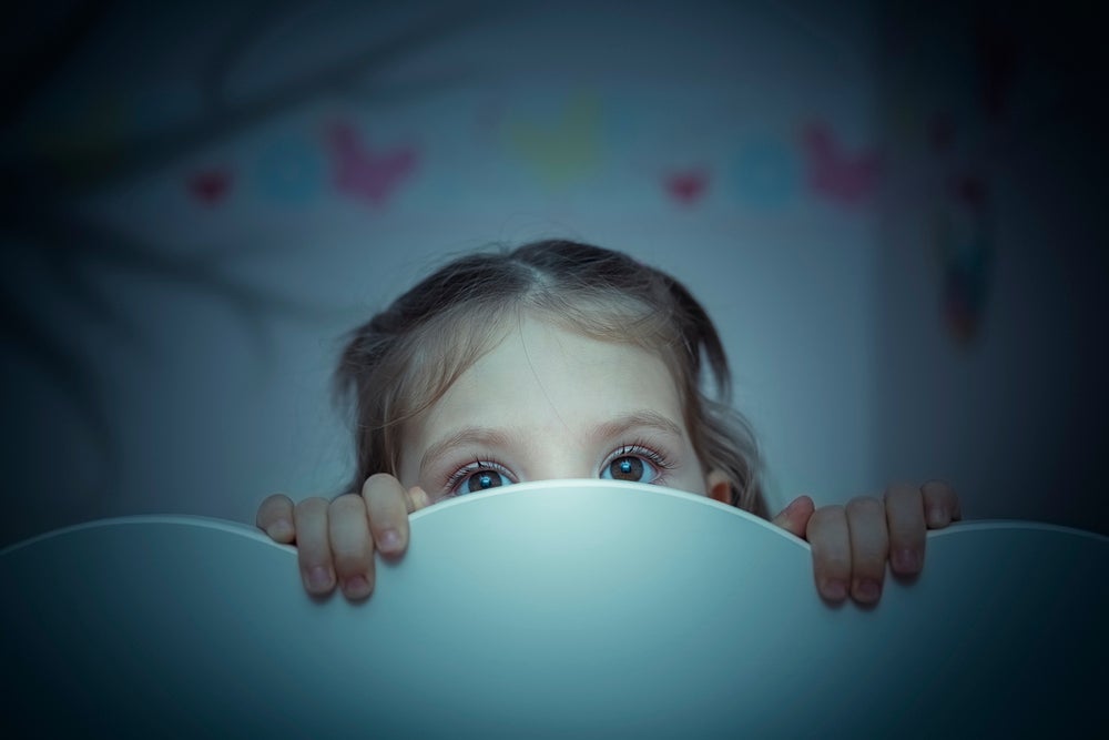 criança se esconde atrás de cabeceira de cama em quarto infantil, rosto com expressão assustada.