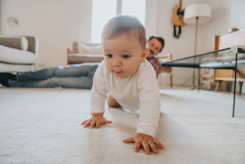 Bebê engatinhando no chão da sala de casa, com a supervisão do pai olhando do canto da sala
