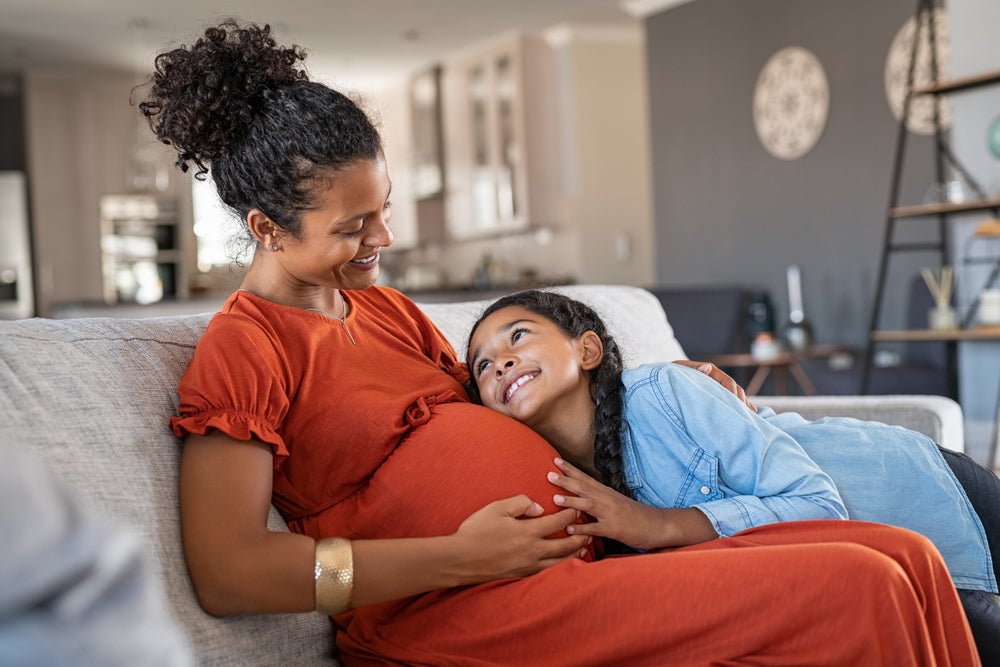 Mãe grávida e filha abraçada em sua barriga, ambas se olham e sorriem, sentadas no sofá da sala de casa.