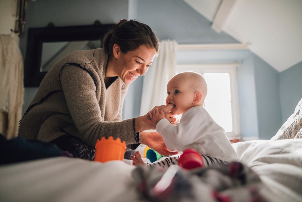 Um bebê está sentado na cama, mordendo o dedo da sua mãe que está na sua boca. A imagem remete ao tema de cárie formada pelo uso da mamadeira