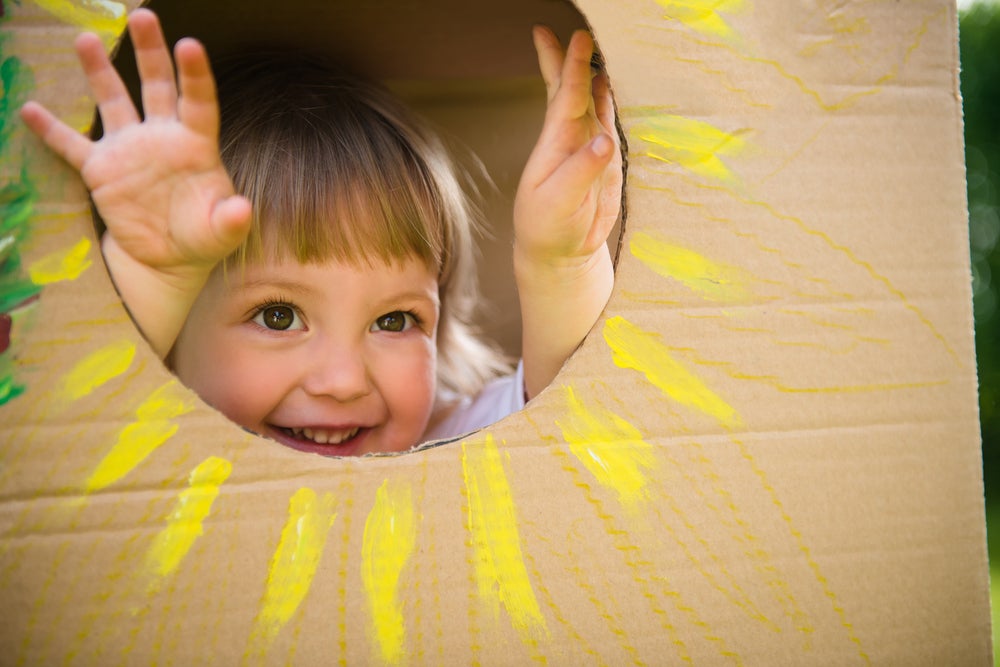 criança sorrindo dentro de uma caixa de papelão, seu rostinho está na parte interior da caixa, num recorte circular onde foi desenhado um sol