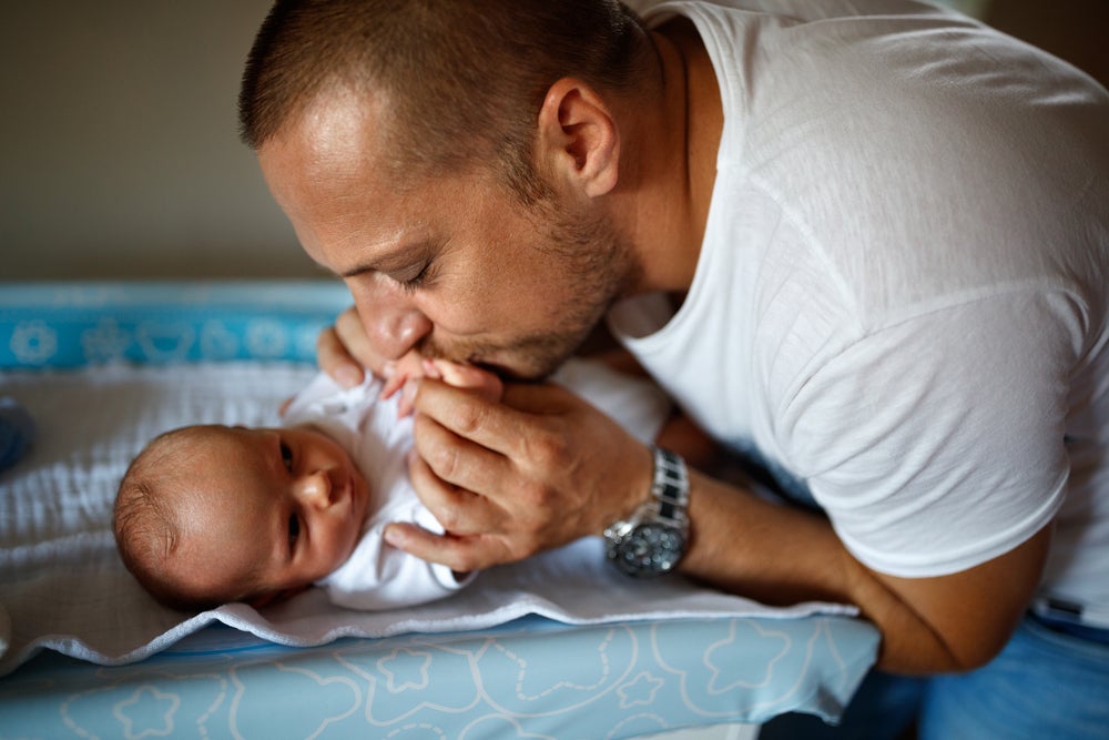 Bebê deitado na cama de barriga para cima com seu pai o acariciando e beijando nos seus pezinhos