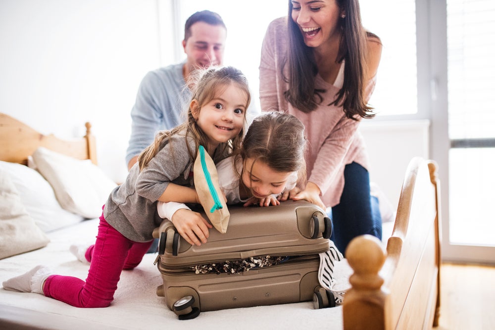 Pai, mãe e duas filhas em cima da cama, tentando fechar uma mala de viagem. Todos estão sorrindo