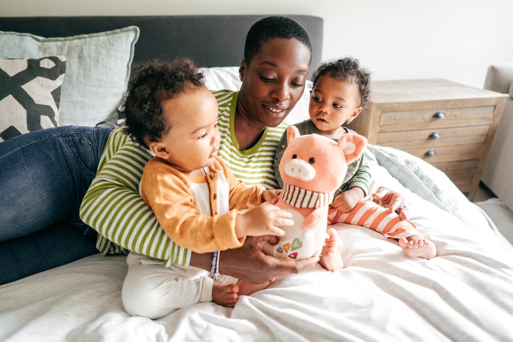Mãe com dois filhos no colo, sentados em cima da cama enquanto brincam com um ursinho de pelúcia