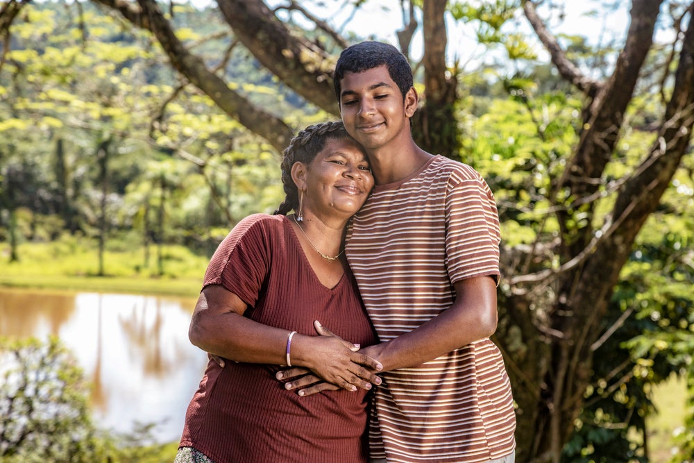 Mulher negra de cabelo preto vestindo uma roupa vermelha abraçando seu filho negro que veste uma roupa listrada com detalhes marrom claro e branco, no fundo um lago e árvores em volta. 