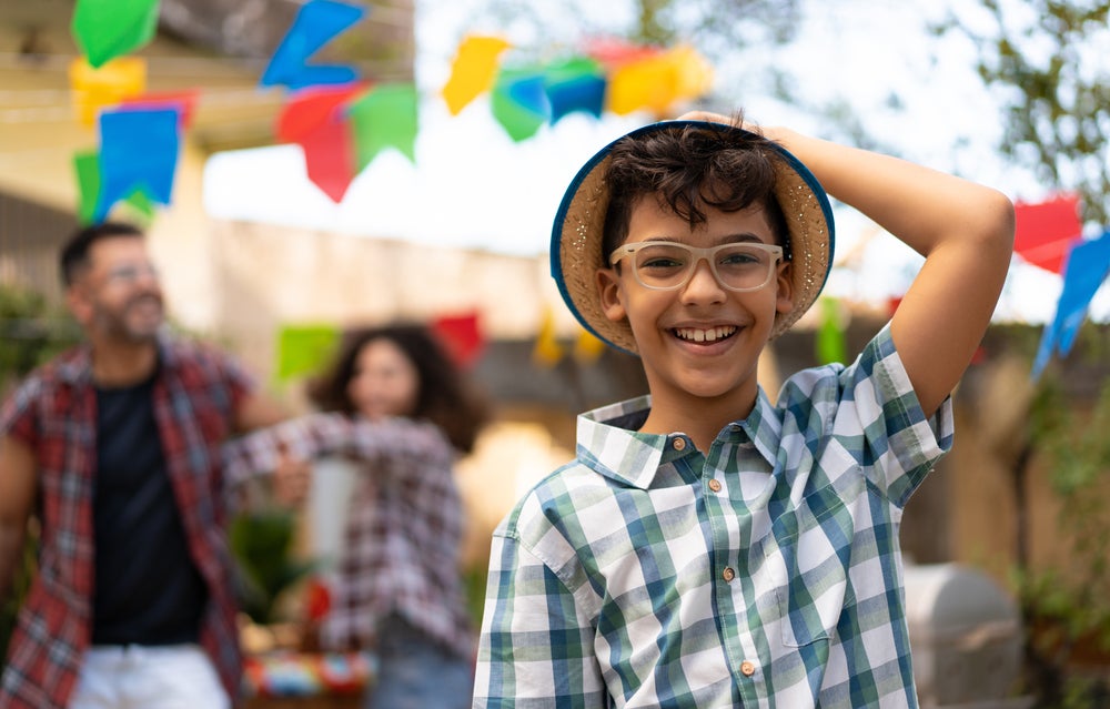 Foto de uma criança sorrindo, usando óculos de grau, camisa listrada e segurando um chapéu de palha. Ao fundo, festa junina, com bandeiras e adultos comemorando.