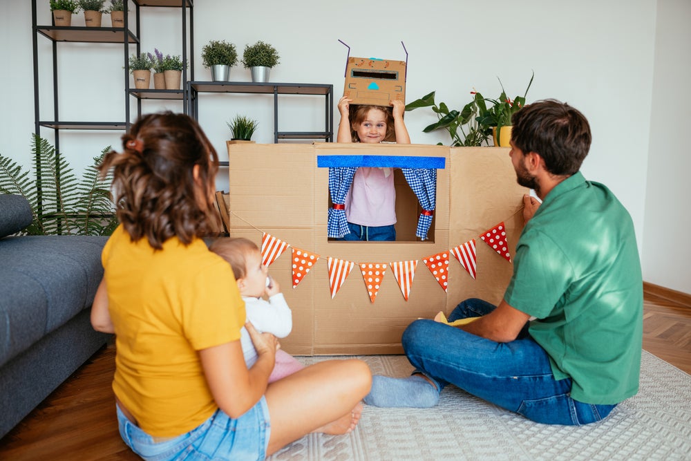 Um homem, uma mulher e um bebê estão sentados no chão de uma sala de estar, de costas, assistindo a uma apresentação de uma menina. Ela está dentro de uma caixa de papelão, simulando uma casinha.