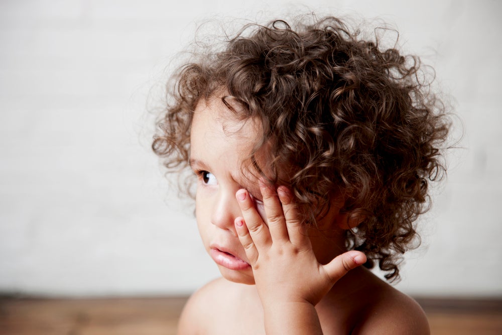 Uma criança, com cabelos cacheados de cor castanha com a mão no rosto, coçando o olho.