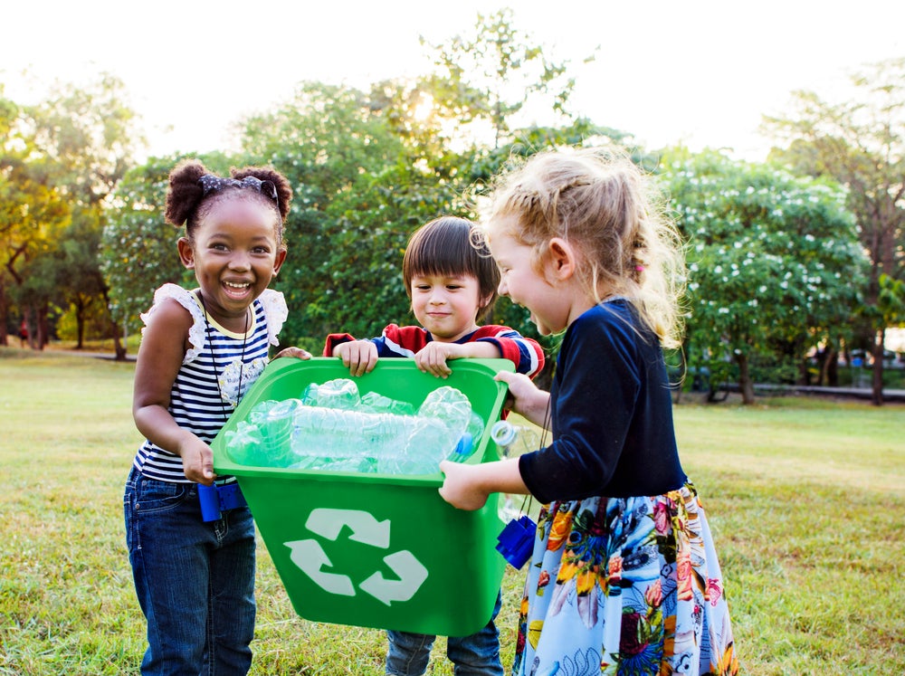 Imagem de três crianças, da esquerda para direita, uma menina, um menino e mais uma menina segurando um lixo com garrafas para reciclagem. Ao fundo, árvores e gramado.