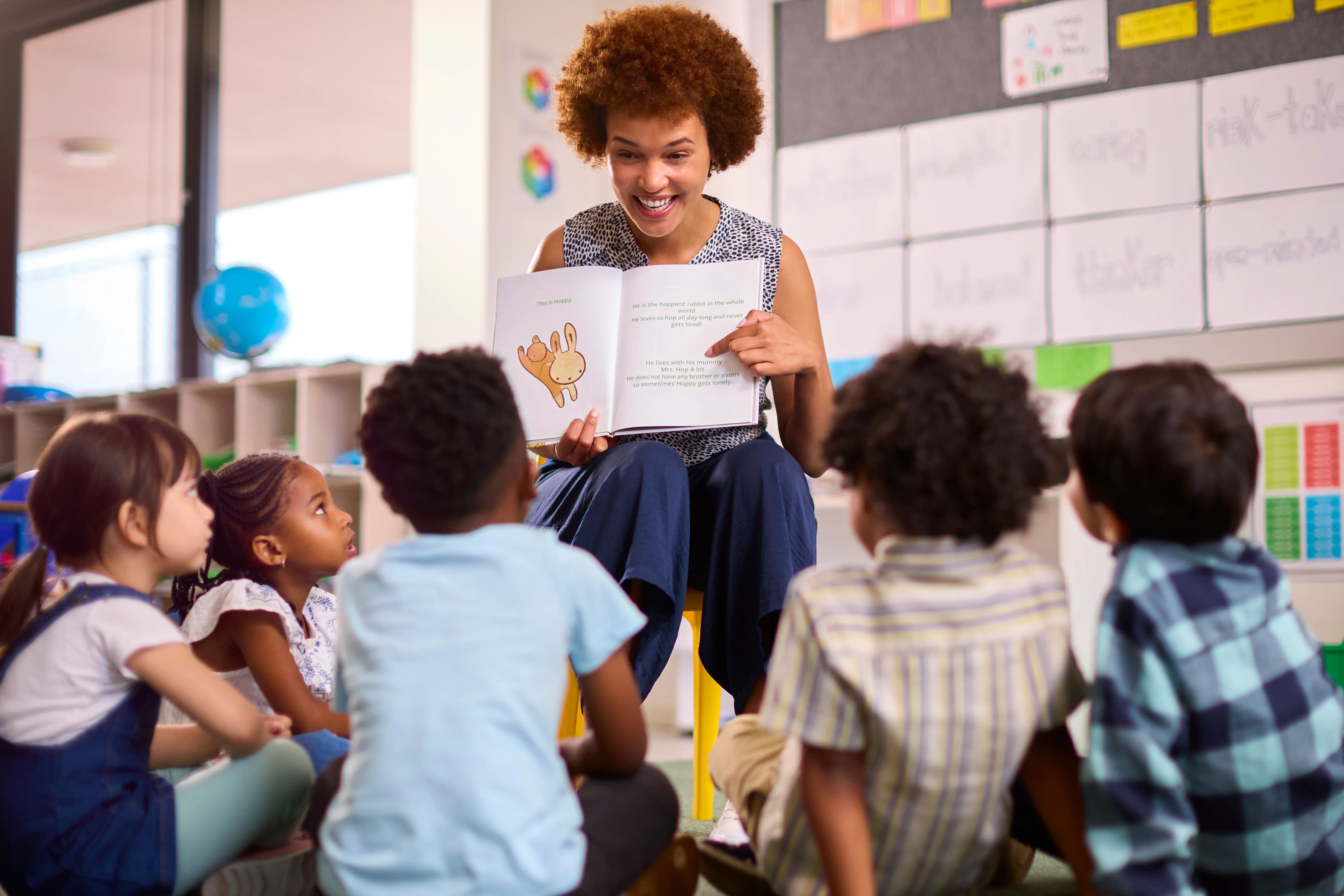 Descrição imagem: Foto de uma mulher segurando um livro e apresentando para as crianças em uma sala de aula sobre educação bilíngue.