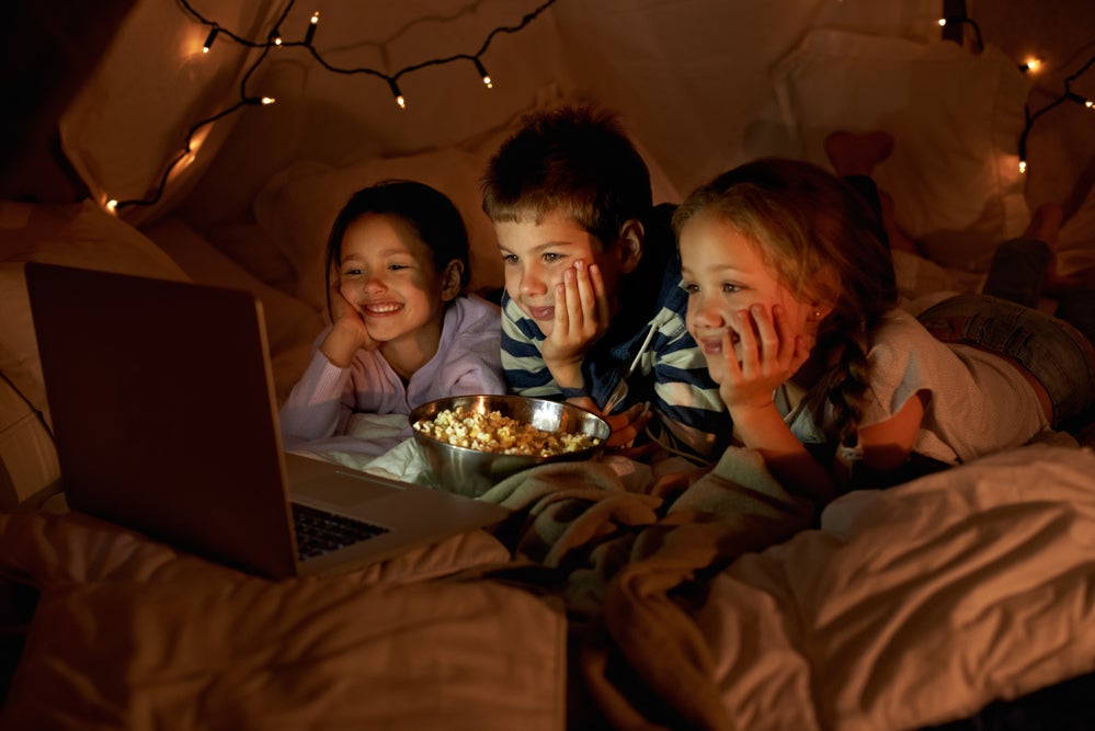 Três crianças, sendo um menino e duas meninas, deitados assistindo no notebook enquanto comem pipoca. Ao fundo, luminárias em uma cabana.