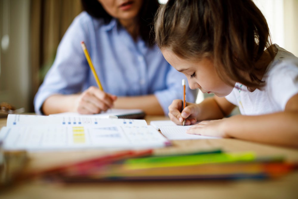 Sentados à mesa, estão uma mulher, que veste uma camisa azul, e uma menina, que veste uma camisa branca. A mulher e a menina estão escrevendo em cadernos, e a menina aparenta estar concentrada. Na mesa, estão alguns livros e canetas coloridas. 