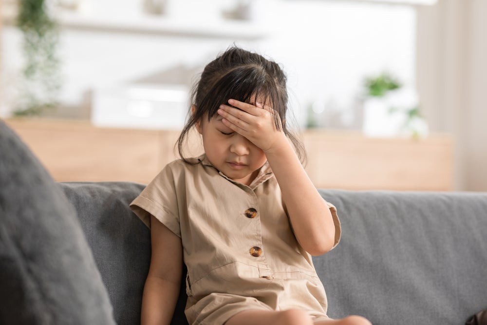 Dentro de uma casa, está uma menina sentada em um sofá cinza, usando um vestido marrom. A menina está com expressão de tristeza, com os olhos fechados e a mão apoiada na cabeça.