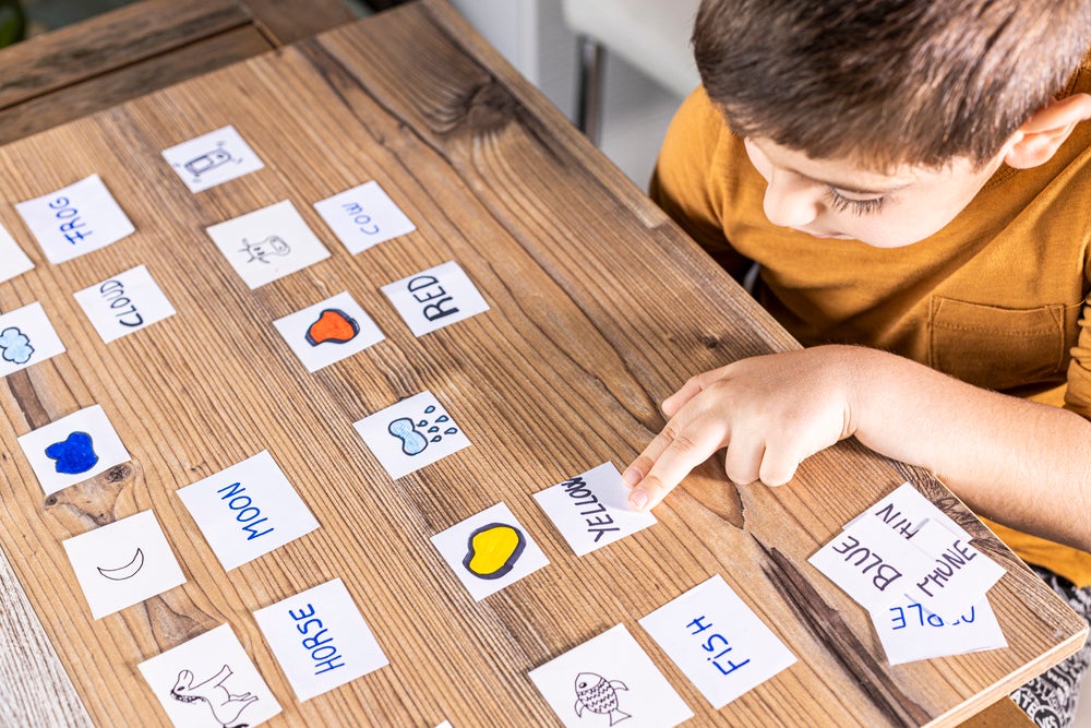 Sentado à mesa, está um menino vestindo uma blusa amarela. Sobre a mesa, há várias cartas com imagens de cores e animais, juntamente com seus respectivos nomes. O menino está associando os nomes às imagens correspondentes.