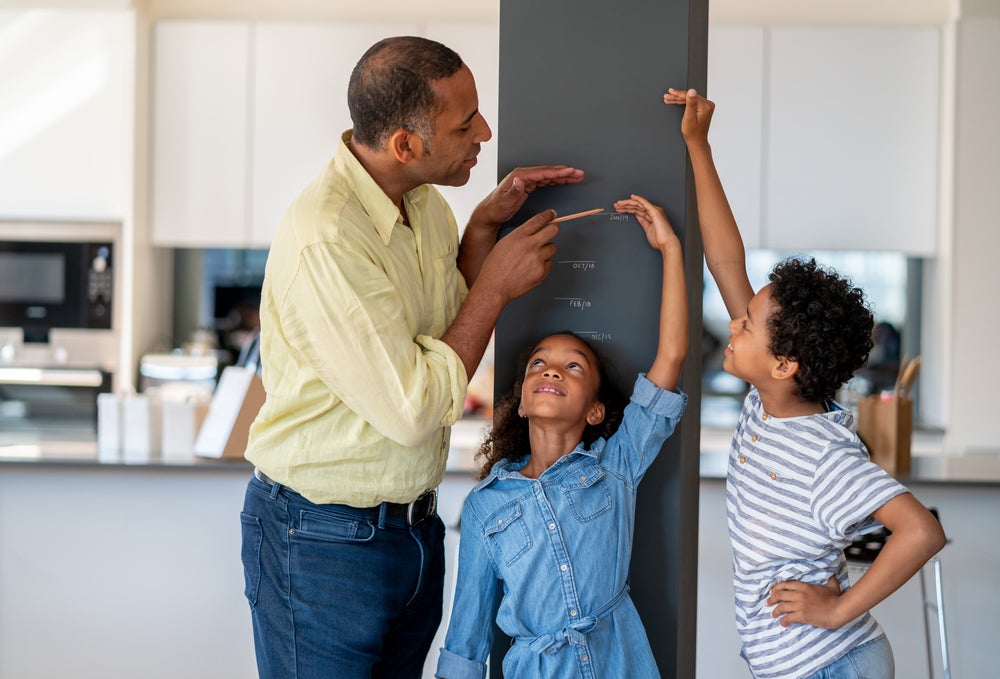 Em uma cozinha, está um homem, uma menina e um menino. A menina está encostada em uma parede, enquanto o homem e o menino marcam na parede qual é a altura dela.