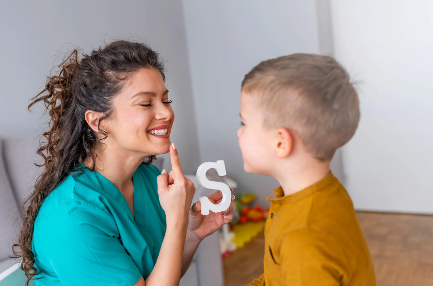 Uma mulher sorrindo, apontando para a própria boca e segurando um objeto em forma de letra “S”, enquanto uma criança observa e tenta imitar a ação da mulher.