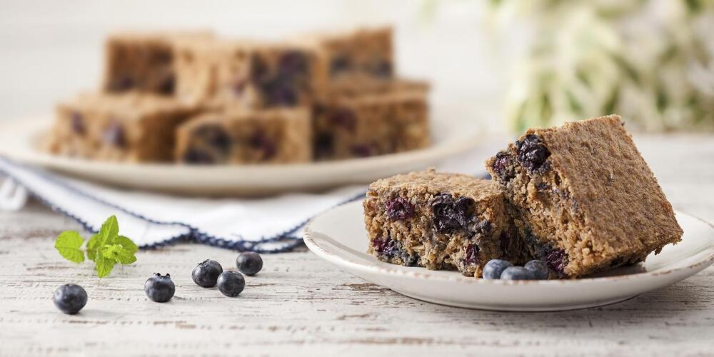 Mesa com dois pratos e alguns bolinhos em formato quadrado em cima dos pratos no sabor de banana e blueberry sem açúcar e sem lactose