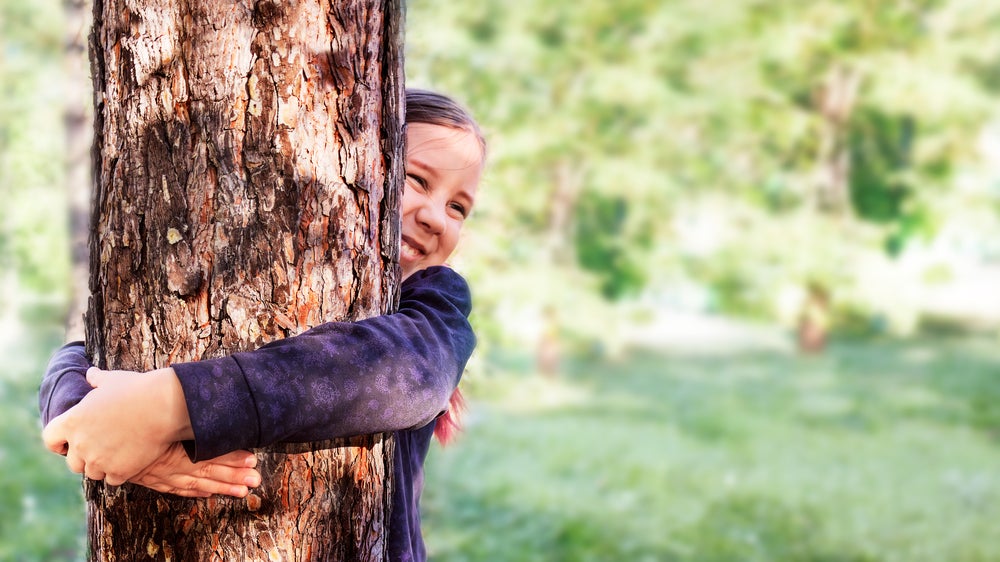  Uma menina de cabelos presos e casaco roxo sorri abraçando uma árvore.