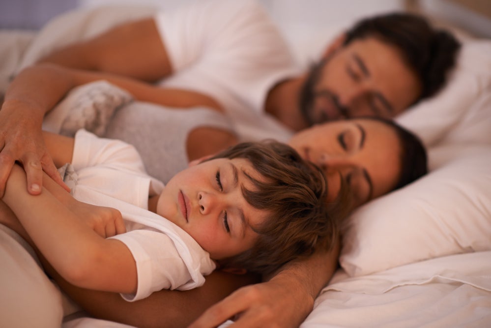 Criança dormindo na cama dos pais. A criança está em primeiro plano, a mãe em segundo e o pai em terceiro