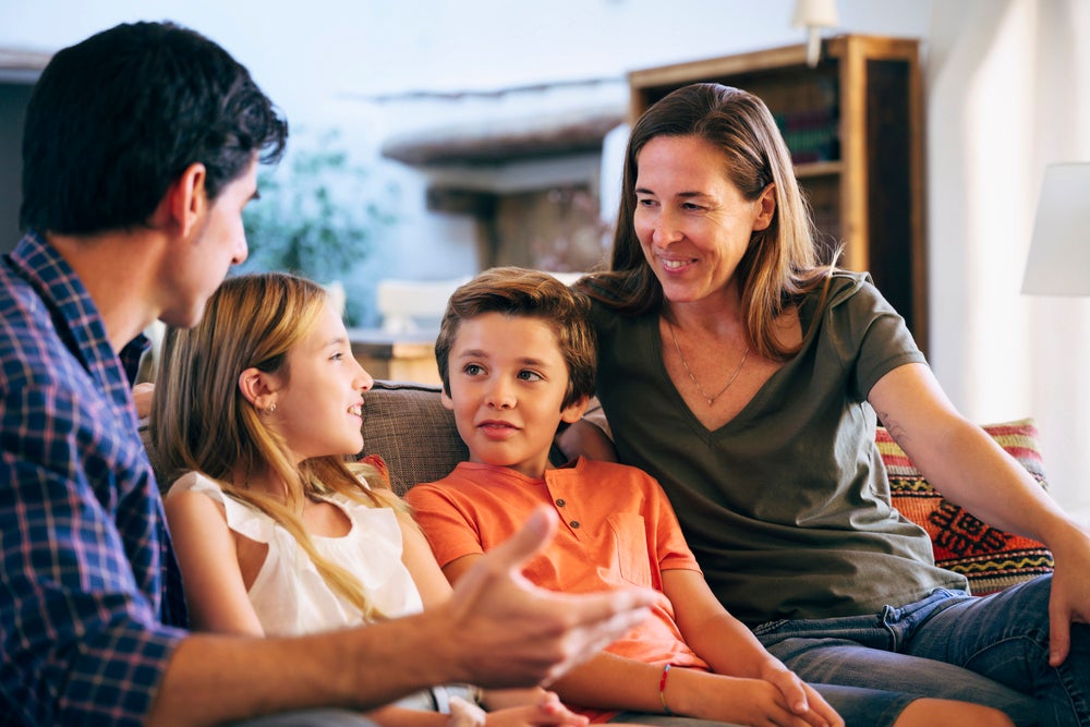 Pai e mãe em um momento de diálogo com filho e filha. Sentados no sofá, o registro ilustra o incentivo da inteligência emocional vindo da família. 