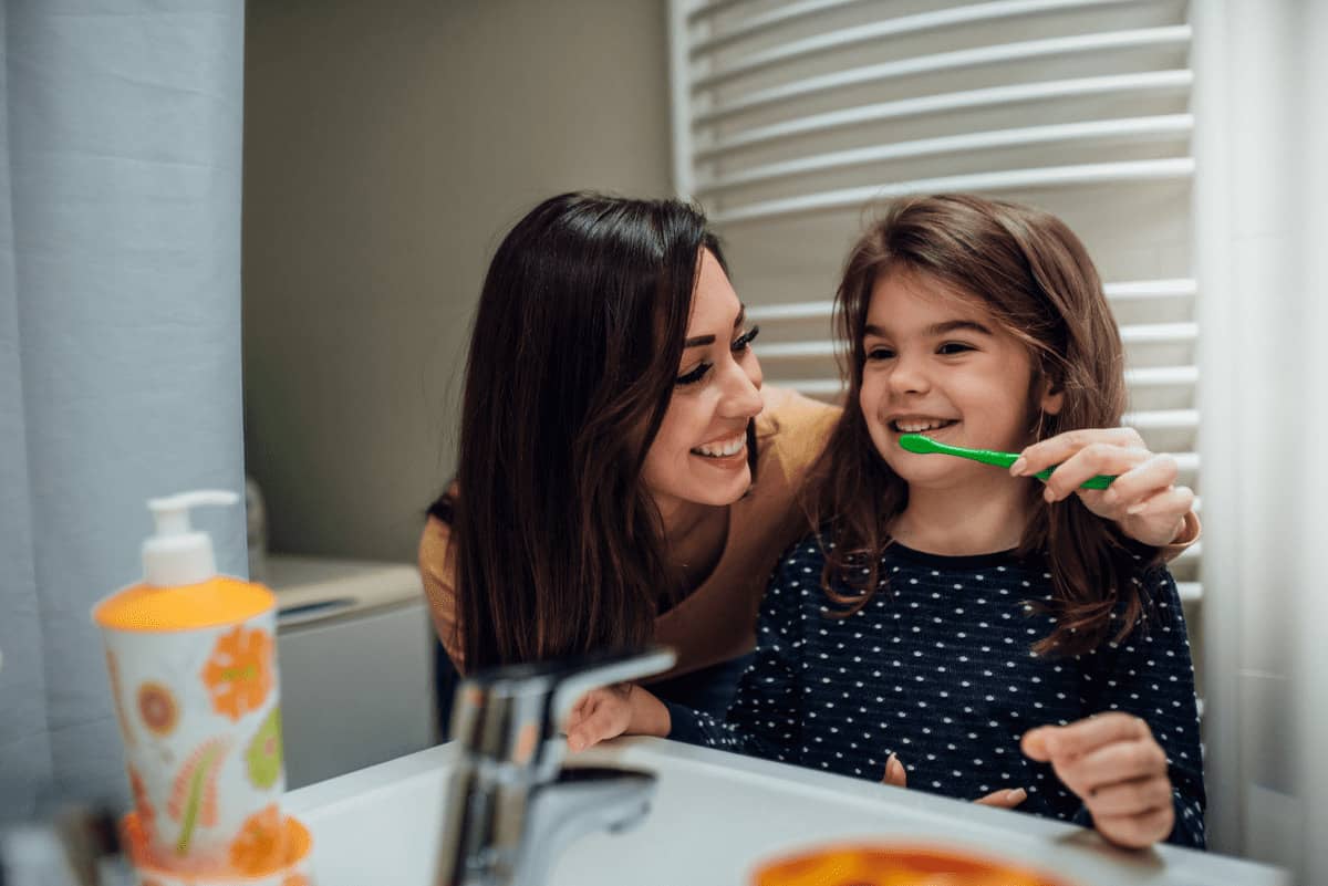 Mãe ajuda sua filha, uma criança de 6 anos, a escovar os dentes em frente ao espelho. Ela sorri e olha para a jovem. 