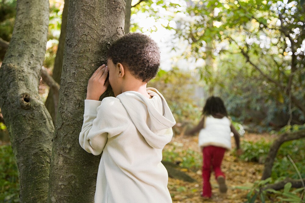 Criança brincando de esconde-esconde, contando encostado numa árvore, enquanto outra criança corre para se esconder