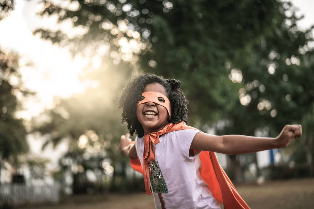 Menina vestida de super-heroína utilizando capa e máscara ao redor dos olhos e se divertindo com a brincadeira para criança. 