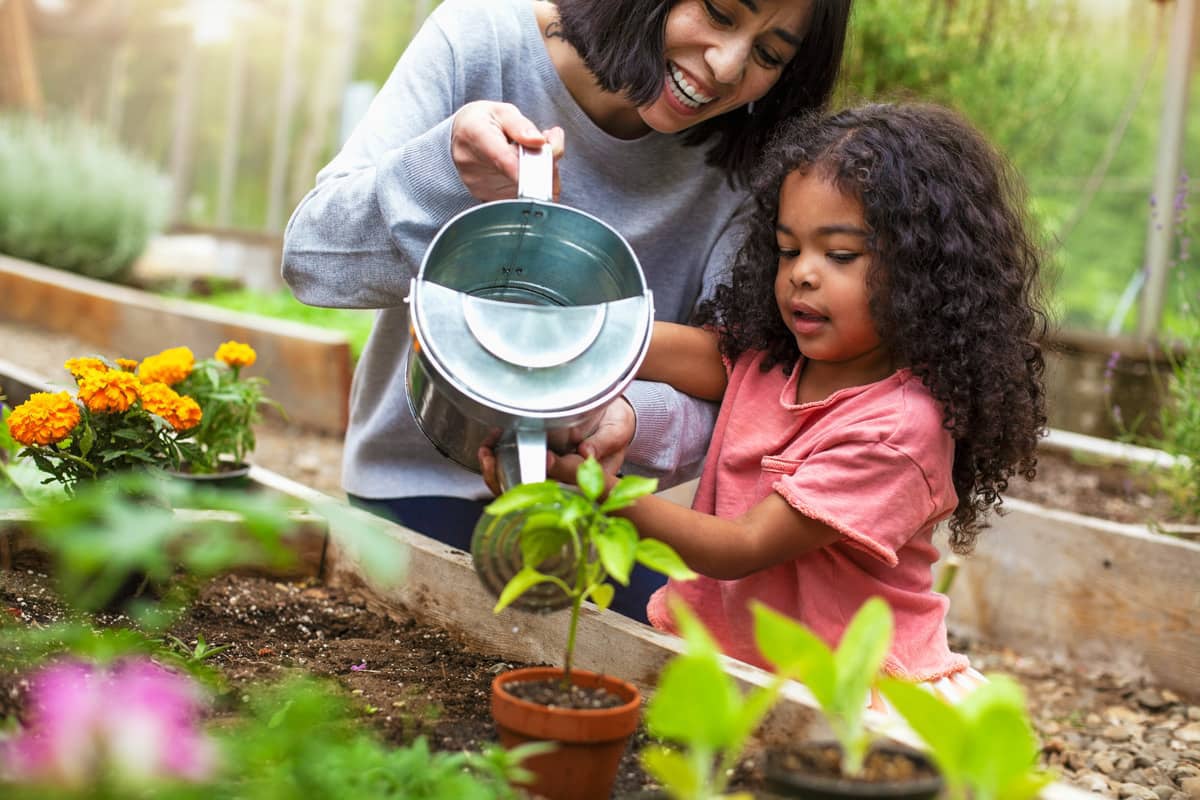 Mãe e filha regam uma planta na horta utilizando, ambas, o mesmo regador.
